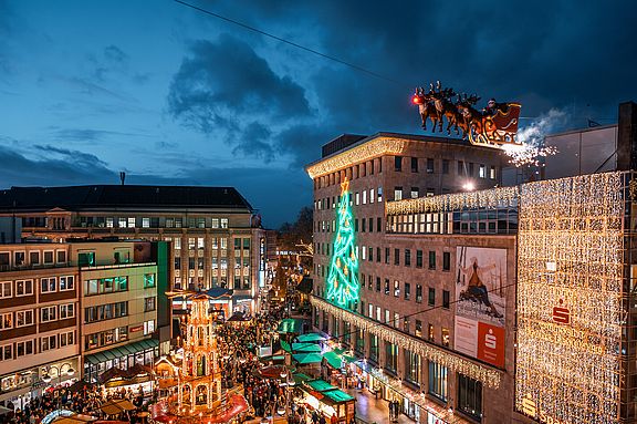Ein lebhafter Weihnachtsmarkt in der Abenddämmerung mit festlichen Lichtern, einem großen geschmückten Baum, Ständen und einer Vorführung mit einem fliegenden Weihnachtsmannschlitten über den Menschenmassen auf einem Stadtplatz, umgeben von beleuchteten Gebäuden.