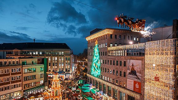 Ein lebhafter Weihnachtsmarkt in der Abenddämmerung mit festlichen Lichtern, einem großen geschmückten Baum, Ständen und einer Vorführung mit einem fliegenden Weihnachtsmannschlitten über den Menschenmassen auf einem Stadtplatz, umgeben von beleuchteten Gebäuden.