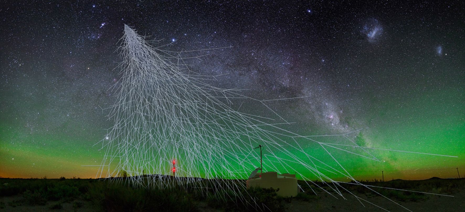 Illustration von kosmischen Strahlen, die auf einen Nachthimmel herabregnen, wobei die Milchstraße und ein grünes Polarlicht sichtbar sind. Im Vordergrund sind ein kleines Observatorium und eine Antenne zu sehen, umgeben von Wüstenlandschaft.