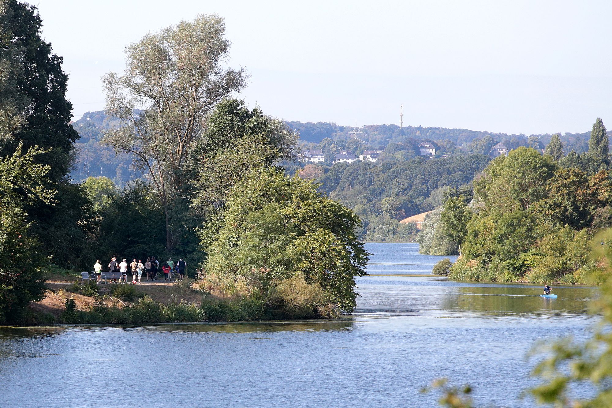 Ein ruhiger Fluss, gesäumt von üppigen Bäumen, mit Menschen, die links auf einem Weg spazieren gehen, und einem einzelnen Kajakfahrer rechts auf dem Wasser. Im Hintergrund sind unter einem hellblauen Himmel Hügel und Häuser zu sehen.