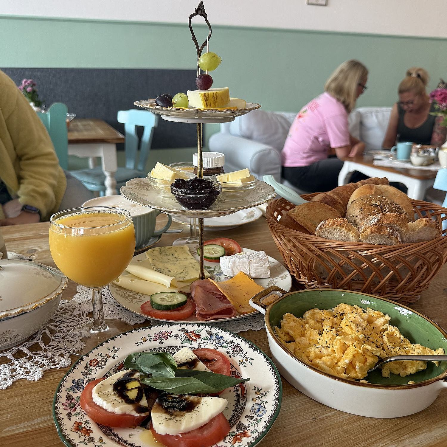 Auf einem Holztisch steht ein Frühstücksbuffet mit Rührei, Tomatenscheiben mit Mozzarella und Basilikum, verschiedenen Käsesorten, Aufschnitt, Brötchen in einem Korb, einem Glas Orangensaft und einem Tablett mit Obst und Gebäck.
