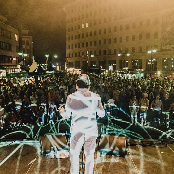 Bochum Musiksommer 2016 Konzert Ein Künstler im weißen Anzug steht bei einem Open-Air-Konzert auf der Bühne vor einer großen Menschenmenge. Die Bühnenbeleuchtung erzeugt dynamische Muster auf dem Boden. Im Hintergrund sind Gebäude und Straßenlaternen zu sehen, die die lebendige Atmosphäre verstärken.