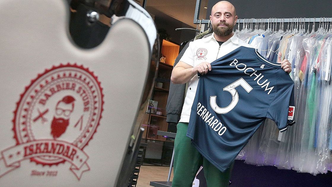Barbier Iskandar Ein Mann steht in einem Geschäft und hält ein blaues Fußballtrikot des VfL Bochum mit dem Namen „Bernardo“ und der Nummer 5 in der Hand. Im Vordergrund steht ein Stuhl mit dem roten Logo „BARBER SHOP ISKANDAR“. Im Hintergrund hängt Kleidung.