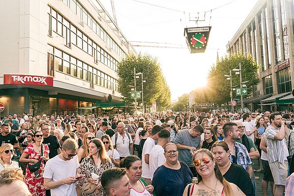 Eine große Menschenmenge versammelt sich auf einer Stadtstraße im Sonnenlicht, umgeben von modernen Gebäuden, Geschäften und Bäumen. Die meisten blicken nach vorne, und die Atmosphäre wirkt lebendig und festlich.