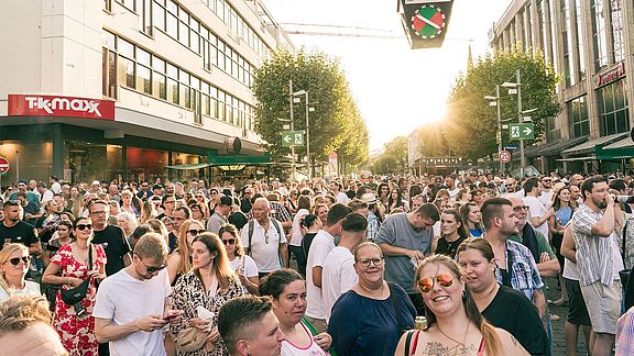 Eine große Menschenmenge versammelt sich auf einer Stadtstraße im Sonnenlicht, umgeben von modernen Gebäuden, Geschäften und Bäumen. Die meisten blicken nach vorne, und die Atmosphäre wirkt lebendig und festlich.