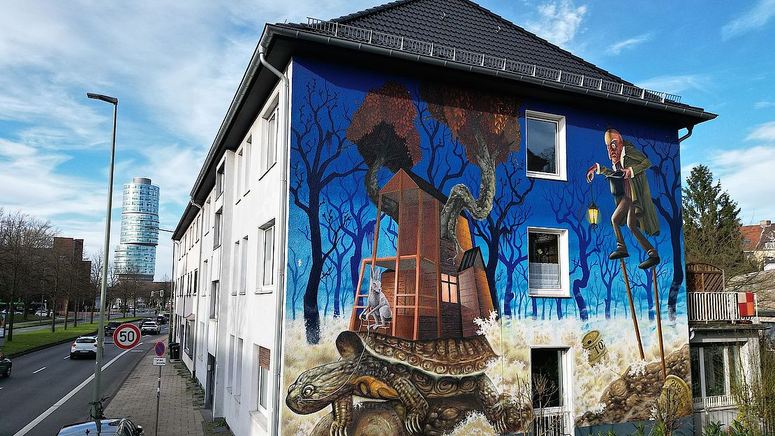 Ein großes Wandgemälde an der Seite eines Gebäudes zeigt eine Schildkröte, die ein kleines Haus in einer surrealen, bewaldeten Landschaft trägt. Ein Mann im Anzug mit einem Vogelkopf steht daneben. Die Szene ist in leuchtenden Blau-, Braun- und Grüntönen unter einem klaren Himmel gehalten.