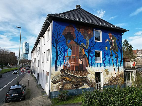 Ein großes Wandgemälde an der Seite eines Gebäudes zeigt eine Schildkröte, die ein kleines Haus in einer surrealen, bewaldeten Landschaft trägt. Ein Mann im Anzug mit einem Vogelkopf steht daneben. Die Szene ist in leuchtenden Blau-, Braun- und Grüntönen unter einem klaren Himmel gehalten.
