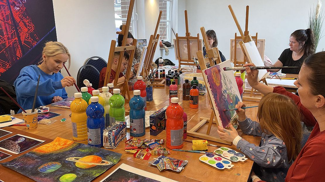 Eine Gruppe von Menschen, darunter auch Kinder, malt in einem hellen Raum farbenfrohe Bilder zum Thema Weltraum. Der Tisch ist mit Farbfläschchen, Pinseln und Staffeleien aus Leinwand bedeckt. Die Atmosphäre ist kreativ und konzentriert.