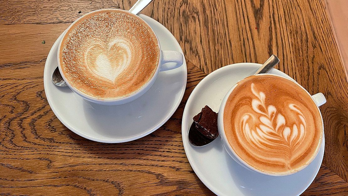 Zwei Cappuccinos mit Latte Art stehen auf einem Holztisch. Die linke Tasse ist mit einem Herzmotiv verziert, während die rechte Tasse ein Blattmuster aufweist. Auf der Untertasse jeder Tasse liegt ein Löffel, daneben ein kleines Stück Schokolade.