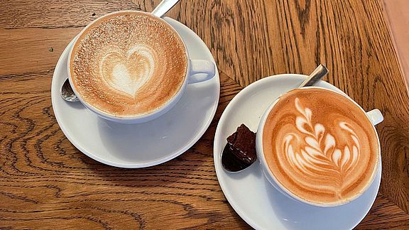 Zwei Cappuccinos mit Latte Art stehen auf einem Holztisch. Die linke Tasse ist mit einem Herzmotiv verziert, während die rechte Tasse ein Blattmuster aufweist. Auf der Untertasse jeder Tasse liegt ein Löffel, daneben ein kleines Stück Schokolade.