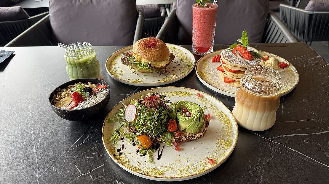 IMG_4326 Ein Tisch mit Tellern mit Avocado-Toast, einem Burger, Pfannkuchen mit Obst, einem Smoothie, einem Eiskaffee, einem Matcha-Getränk und einer Schale mit Obst und Müsli, in einem modernen Restaurant mit grauen Stühlen.
