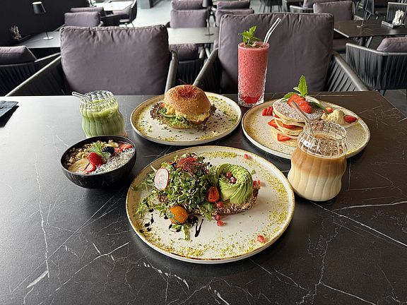 Ein Tisch mit Tellern mit Avocado-Toast, einem Burger, Pfannkuchen mit Obst, einem Smoothie, einem Eiskaffee, einem Matcha-Getränk und einer Schale mit Obst und Müsli, in einem modernen Restaurant mit grauen Stühlen.