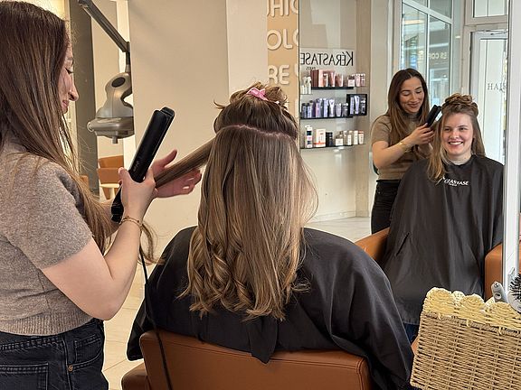 Zwei Frauen lassen sich in einem Friseursalon die Haare stylen. Zwei Friseurinnen arbeiten mit Stylinggeräten an ihren Kundinnen, die lächelnd auf den Stühlen sitzen. Im Hintergrund sind Haarprodukte und die Saloneinrichtung zu sehen.