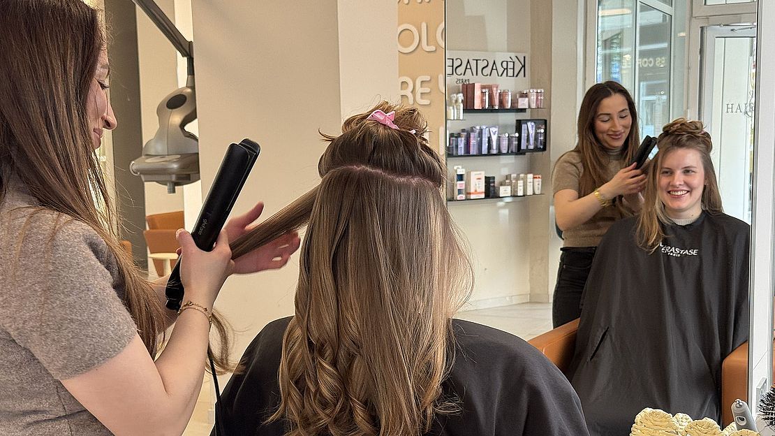 Zwei Frauen lassen sich in einem Friseursalon die Haare stylen. Zwei Friseurinnen arbeiten mit Stylinggeräten an ihren Kundinnen, die lächelnd auf den Stühlen sitzen. Im Hintergrund sind Haarprodukte und die Saloneinrichtung zu sehen.