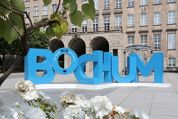 Große blaue Buchstaben mit der Aufschrift „BOCHUM“ stehen prominent auf einem öffentlichen Platz vor einem historischen Steingebäude. Weiße Blumen und grüne Blätter rahmen den Vordergrund ein.