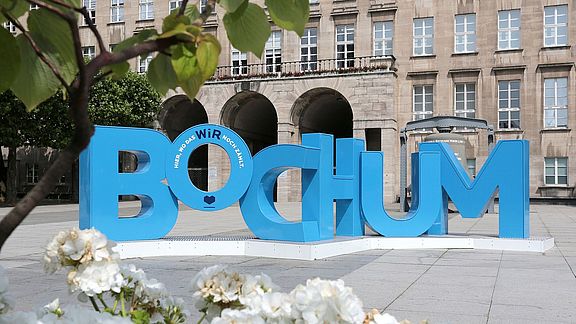 Große blaue Buchstaben mit der Aufschrift „BOCHUM“ stehen prominent auf einem öffentlichen Platz vor einem historischen Steingebäude. Weiße Blumen und grüne Blätter rahmen den Vordergrund ein.