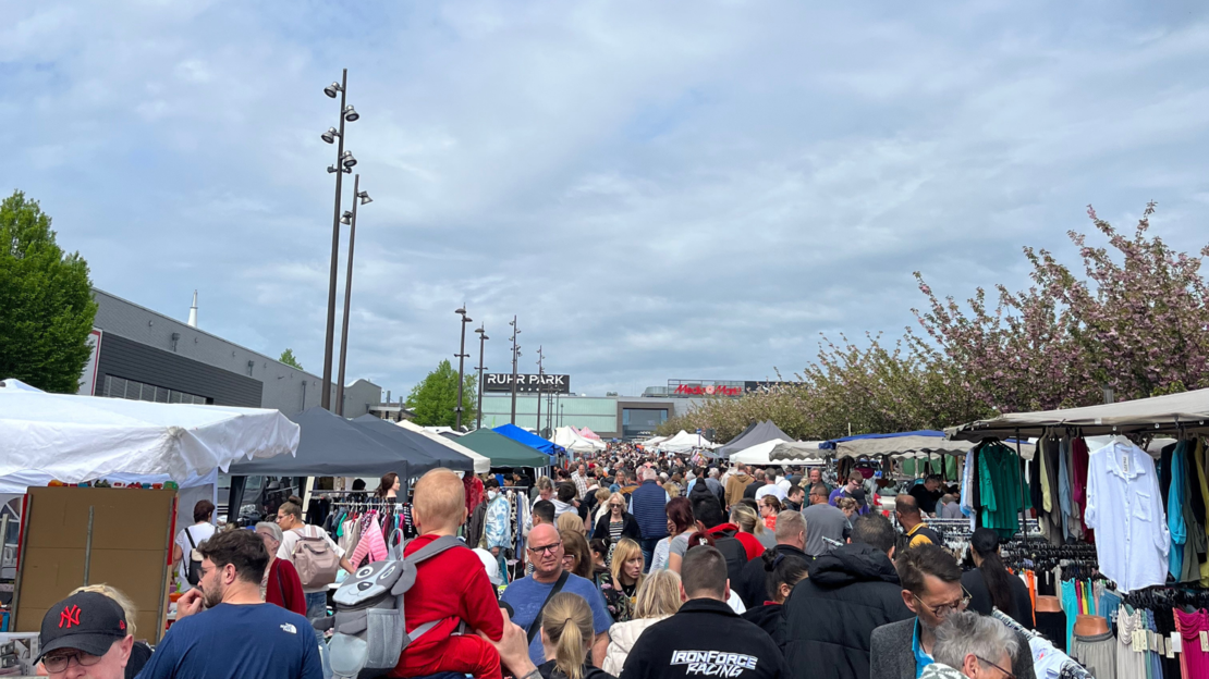 Ein geschäftiger Freiluftmarkt voller Menschen, die zwischen den Ständen umherlaufen. Händler bieten unter weißen Vordächern Kleidung und verschiedene Artikel an. Der Himmel ist bedeckt und blühende Bäume säumen den Marktweg.
