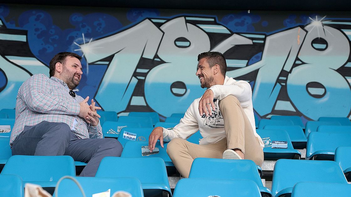Zwei Männer sitzen auf blauen Stadionsitzen und unterhalten sich. Einer trägt ein kariertes Hemd und der andere ein weißes Sweatshirt. Im Hintergrund sind Graffiti mit den Zahlen „1860“ und ein blau-graues Farbschema zu sehen.